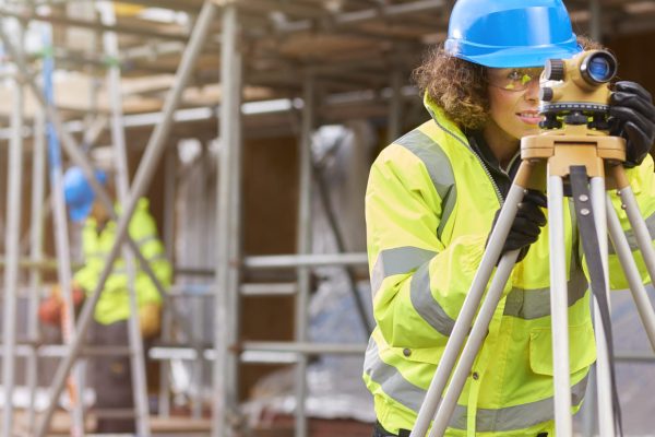 a female construction worker stands behind a builder's level on a  building site . In the background a colleague is working under scaffolding